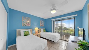 Een hotelkamer met twee bedden, een plafondventilator, een balkon met uitzicht en kunstwerken aan de muur.