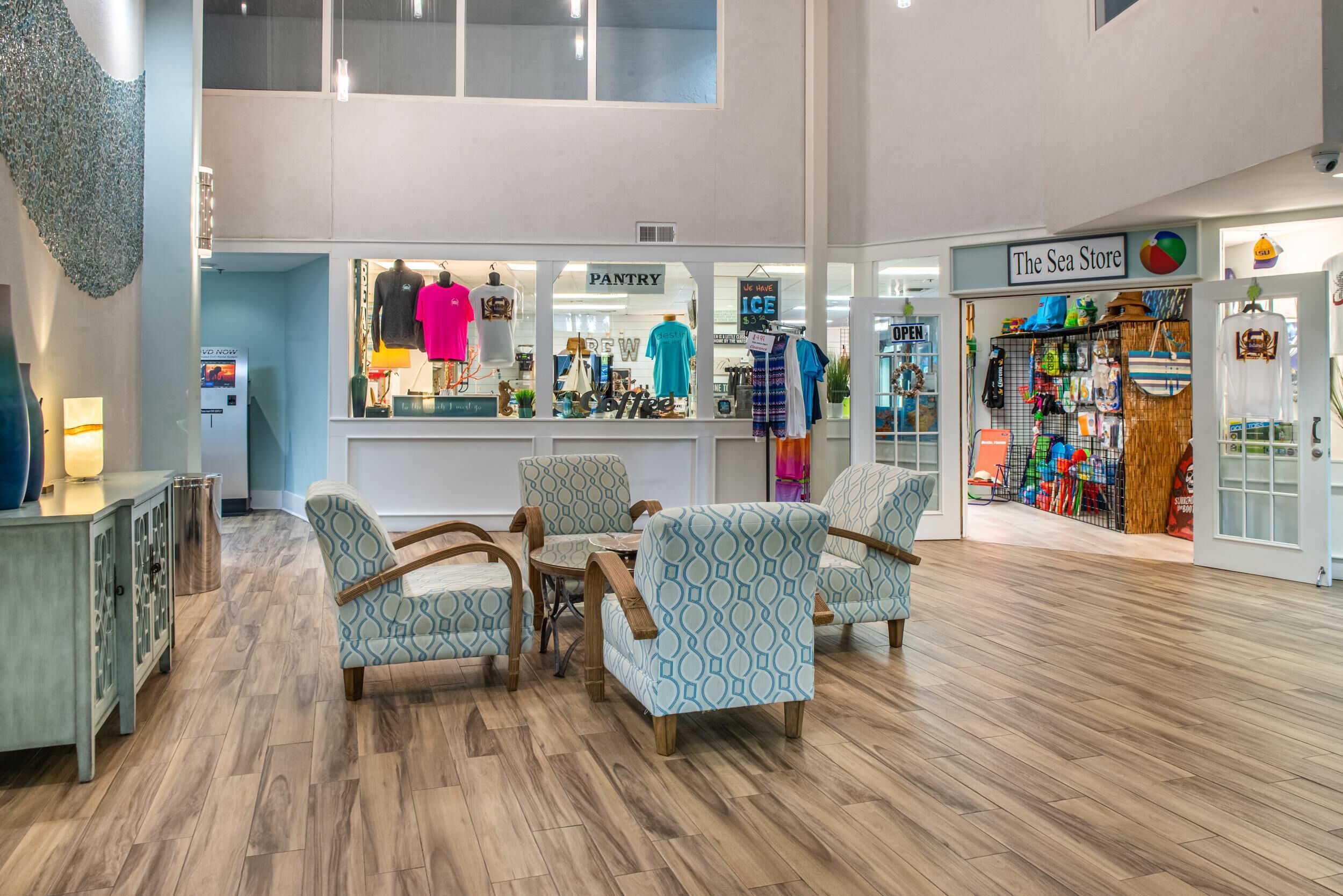 Um hall espaçoso com teto alto, área de estar central com cadeiras estampadas, uma cozinha compacta visível, e uma loja chamada "The Sea Store" com diversos artigos em exposição.