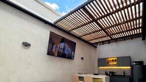 Un área de estar exterior moderna con jacuzzi, televisor en la pared y zona de cocina.