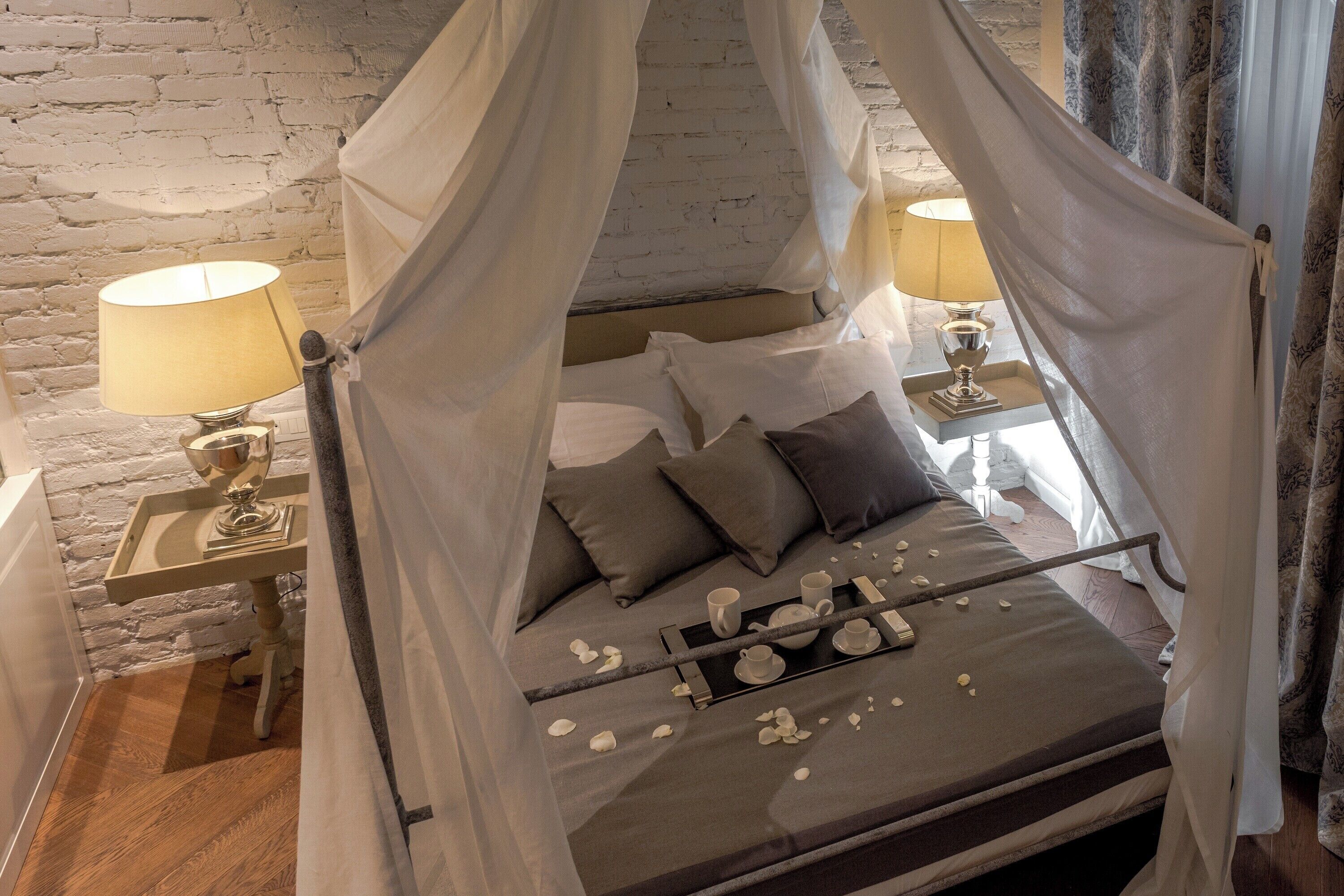 Ein Bett mit Baldachin, Nachttischlampen, ein Tablett mit Tassen und einem Teekessel, sowie eine Backsteinwand im Hintergrund.