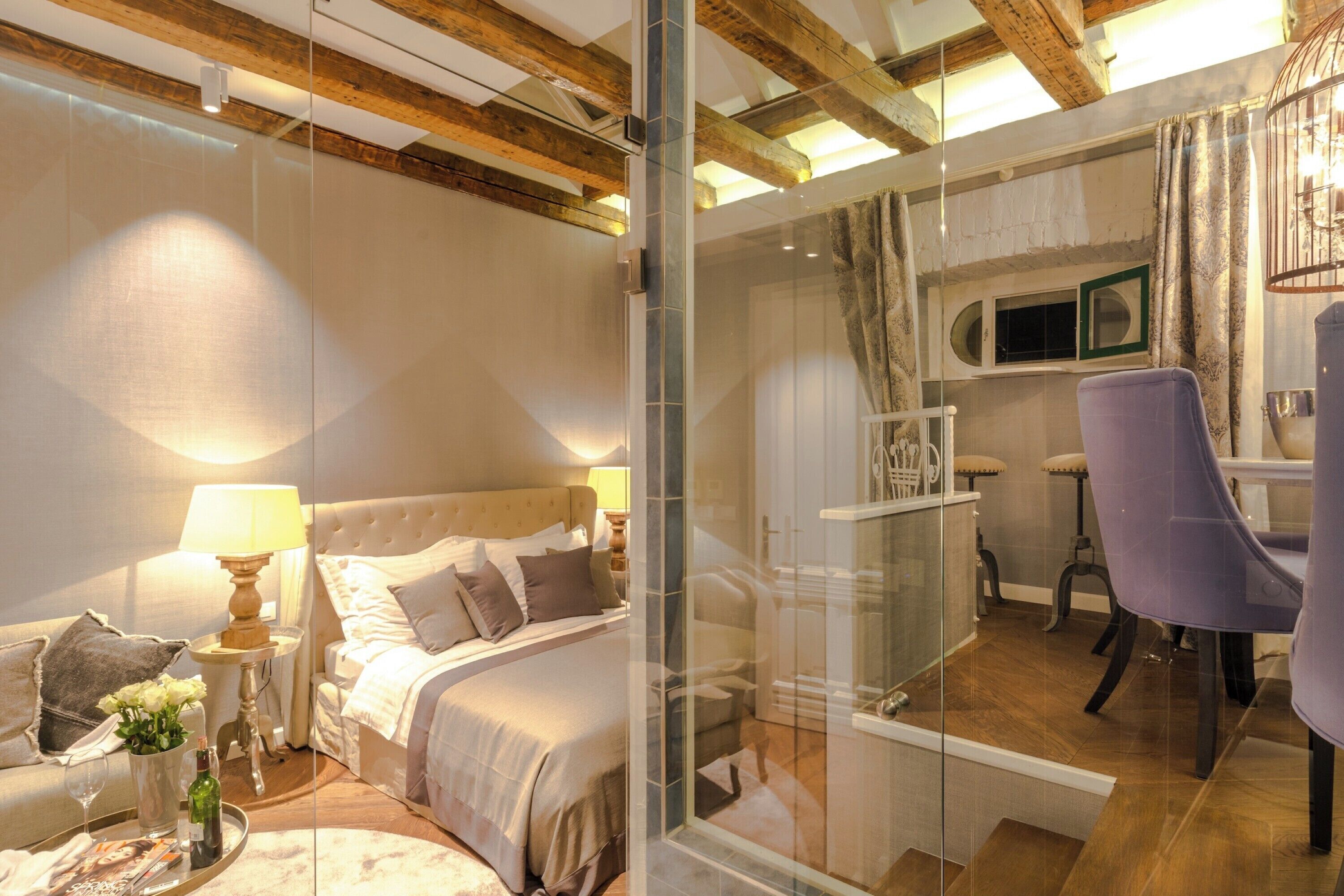 A bedroom with a bed, bedside table, lamp, and a glass partition separating it from a bathroom with a shower and chair.