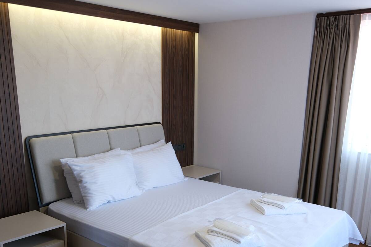 Una cama bien hecha con sábanas y almohadas blancas, una mesita de noche y suelo de madera.