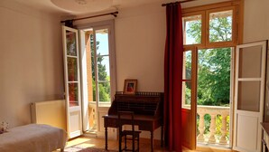 Een slaapkamer met een bed, een bureau, een piano en uitzicht op groen door open ramen.