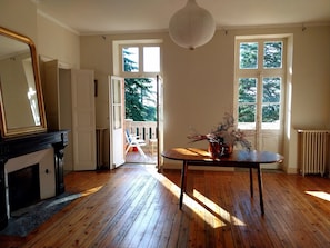 A room with wooden flooring, a fireplace, a table with a vase, and a view through an open door to a balcony. - Le Gite de Douine (Avignonet-Lauragais)