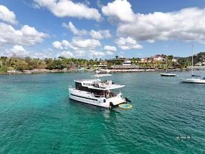Eine weiße Yacht mit abgedecktem Deck und einem kleinen Schlauchboot, vor Anker in klarem türkisblauem Wasser, mit einer Küstenstadt und Gebäuden im Hintergrund.