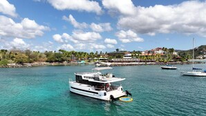 Een jachthaven met een witte boot genaamd STAY-01, kalm turquoise water en een achtergrond van een kustplaatsje met gebouwen en palmbomen.