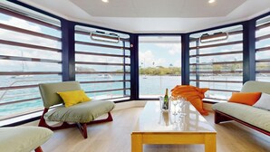 Een modern interieur met een houten vloer, twee banken met kussens, een salontafel en grote ramen met uitzicht op het water.