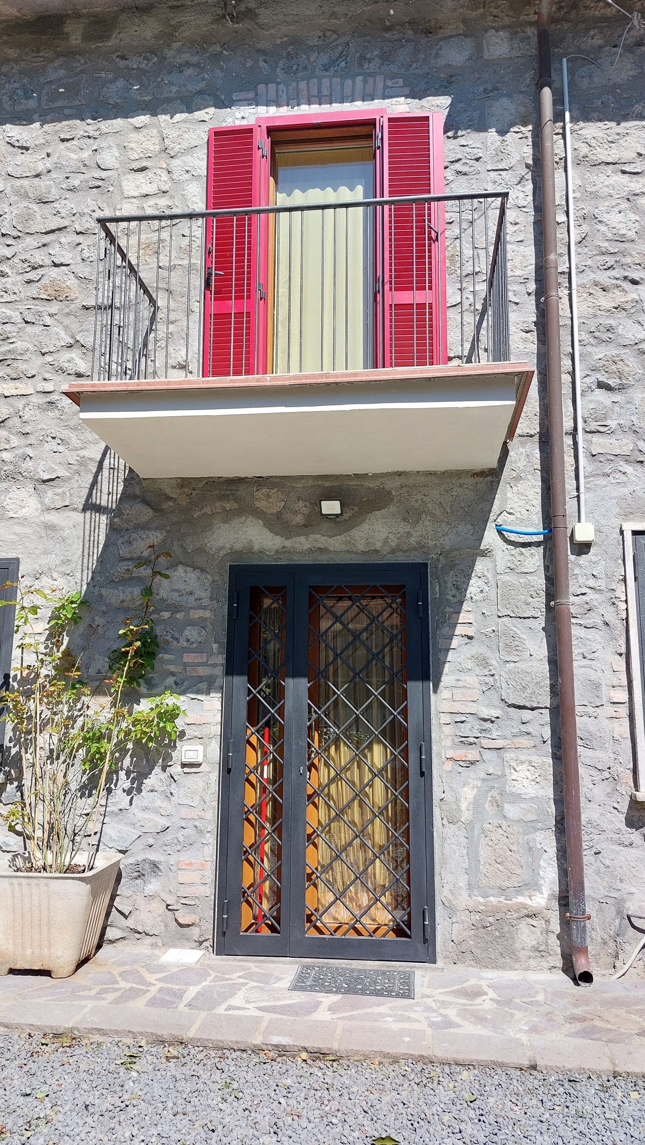 Un edificio con un balcón, una puerta con celosía metálica y un muro de piedra.