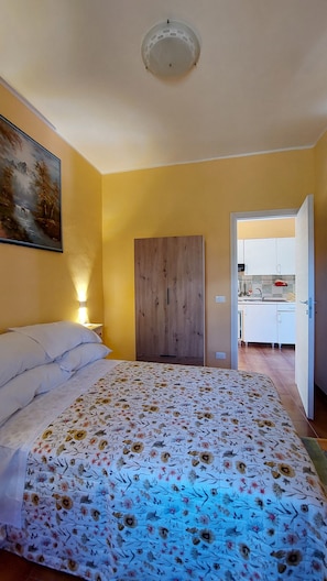 A bedroom with a bed, a painting, a wardrobe, and a view into a kitchen.