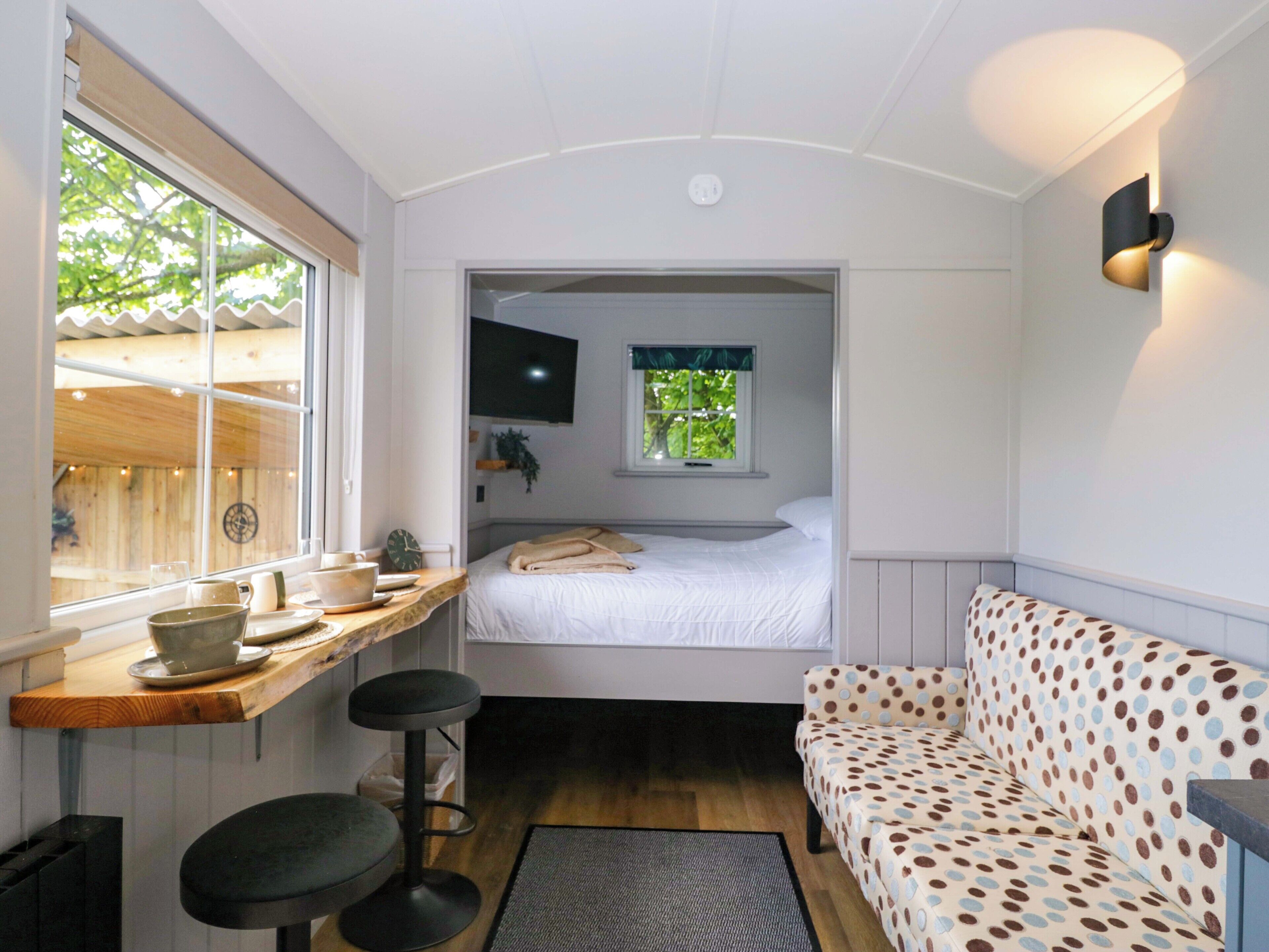 A compact room with a bed, a small table with a cup and saucer, a bar stool, a patterned sofa, and a window with a view of greenery.
