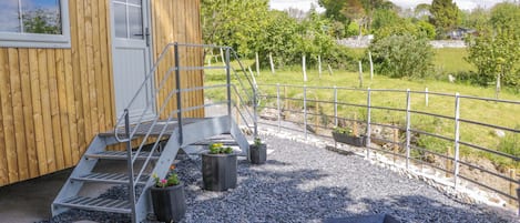 A wooden cabin with a metal staircase leading to a door, a gravel patio with a metal grill, and a metal fence enclosing a grassy area.