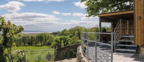 Un muro de piedra con una reja de metal, un edificio de madera con una escalera y vistas a un paisaje verde.