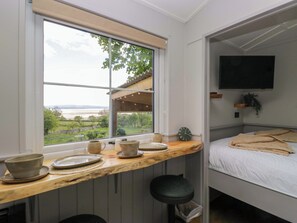 A bedroom with a bed, a TV mounted on the wall, a window with a view of the outdoors, and a small kitchenette area with a countertop and stools. - Little Hut (Grange-over-Sands)