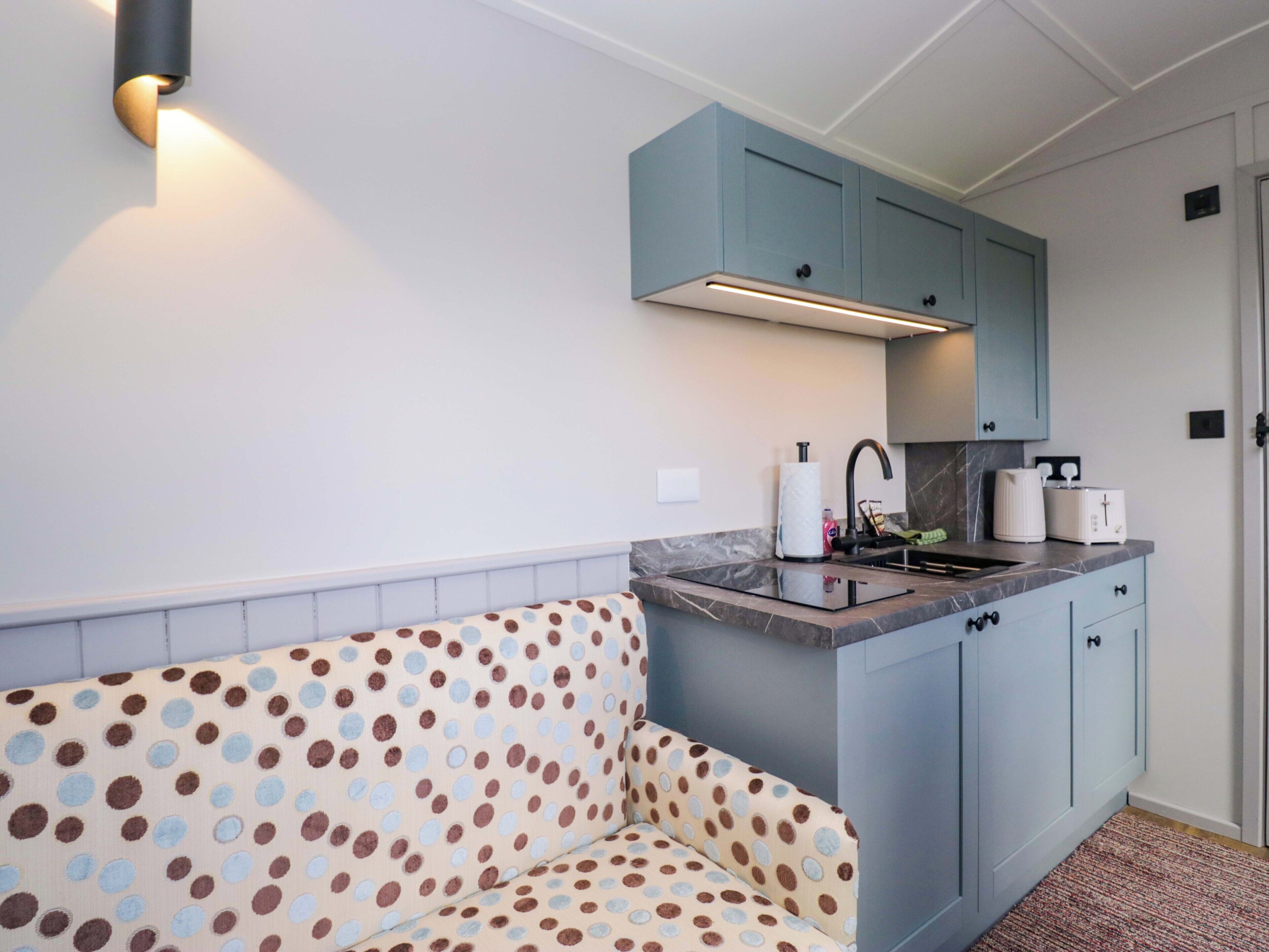 A kitchenette with a built-in stove, a sink, and a countertop. There is a polka-dotted sofa against the wall and a light-colored rug on the floor.