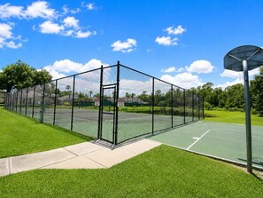Una cancha de tenis con una red, rodeada por una valla, y un aro de baloncesto a la derecha.