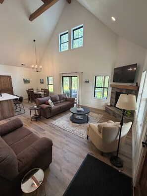A spacious living area with a high ceiling, a fireplace, a television, and a dining area with a table and chairs.