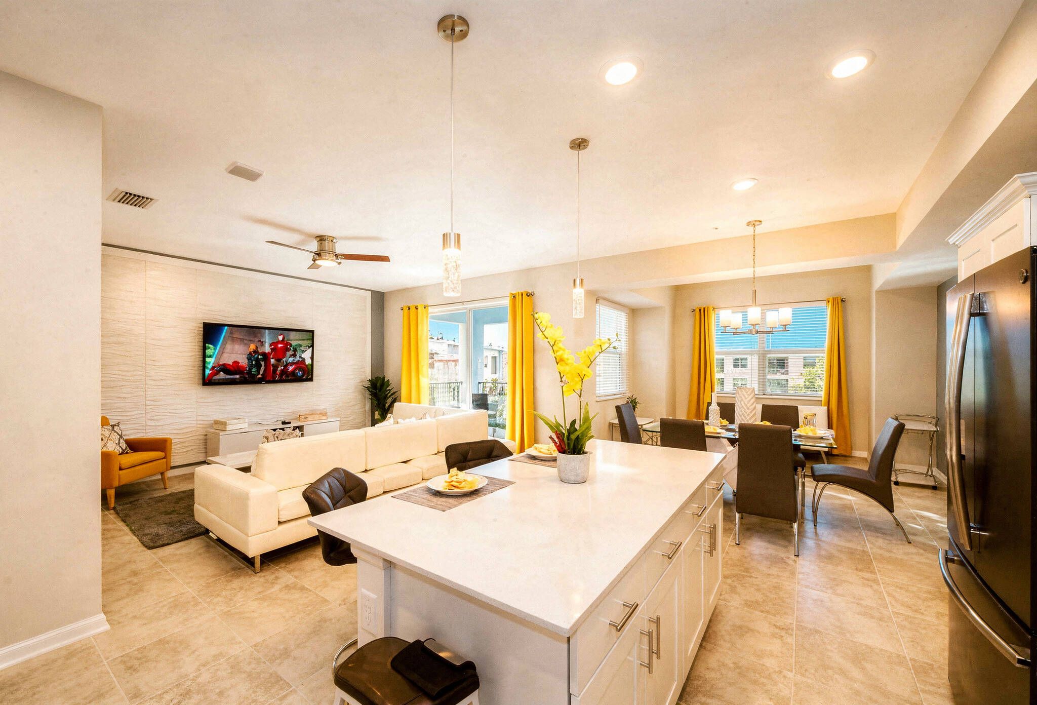 A modern kitchen with a central island, dining area with yellow curtains, and a living room with a sofa and TV.