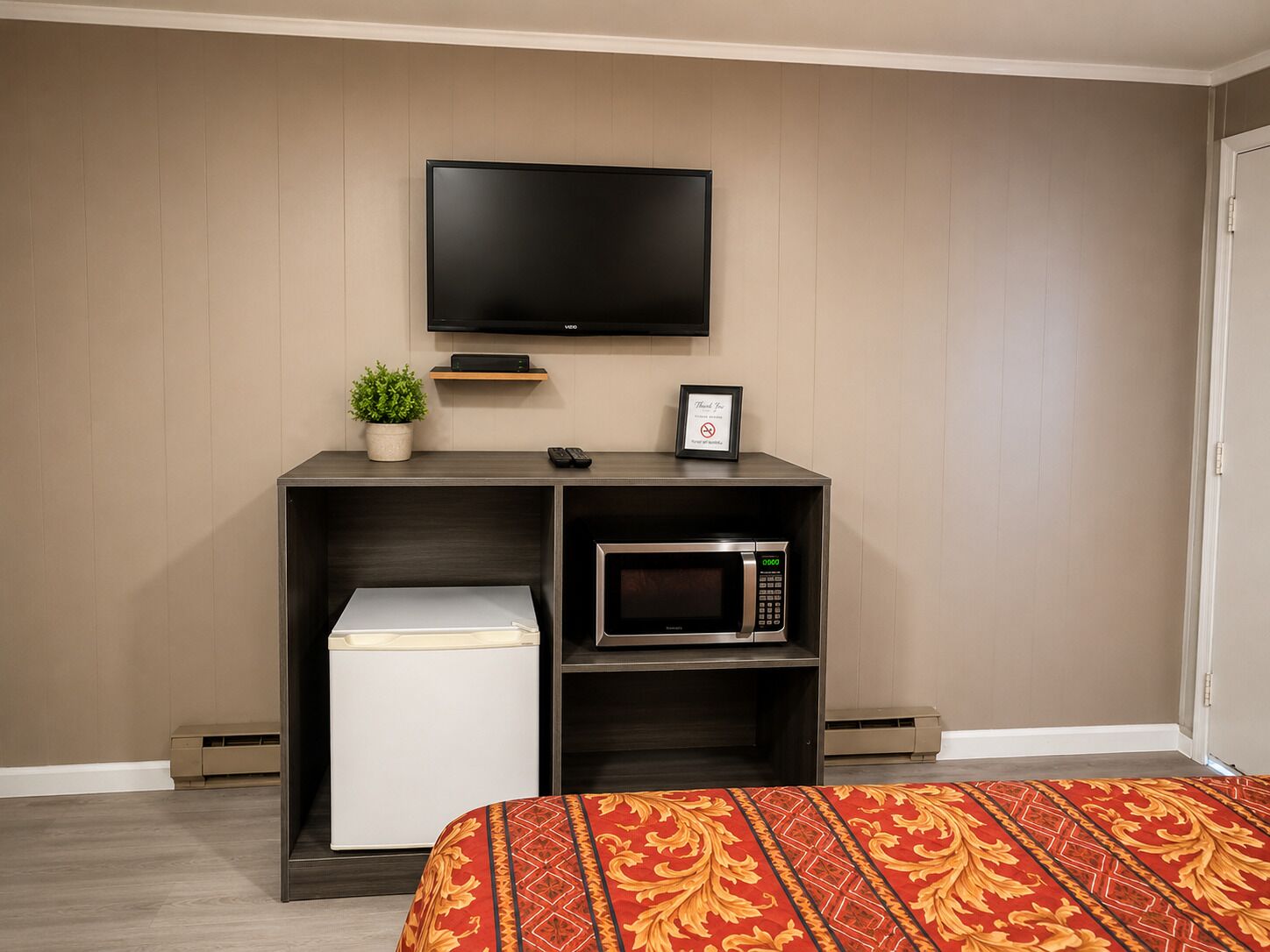 A hotel room with a bed, a TV mounted on the wall, a microwave, a small shelf with a plant, and a framed sign.