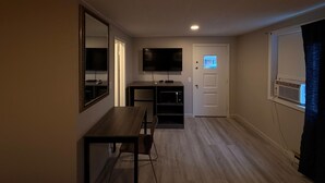 Habitación con televisión de pantalla plana en la pared, un escritorio de madera con silla, una puerta con ventanal y un equipo de aire acondicionado.