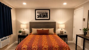 Habitación de hotel con una cama grande, dos mesitas de noche con lámparas, una fotografía enmarcada de un paisaje urbano en la pared, una planta en maceta sobre la mesita auxiliar y una puerta con cerradura.