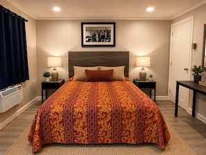 A hotel room with a large bed, two bedside tables with lamps, a framed cityscape picture on the wall, a potted plant on the side table, and a door with a lock. - Patio Court Motel (Quakertown)