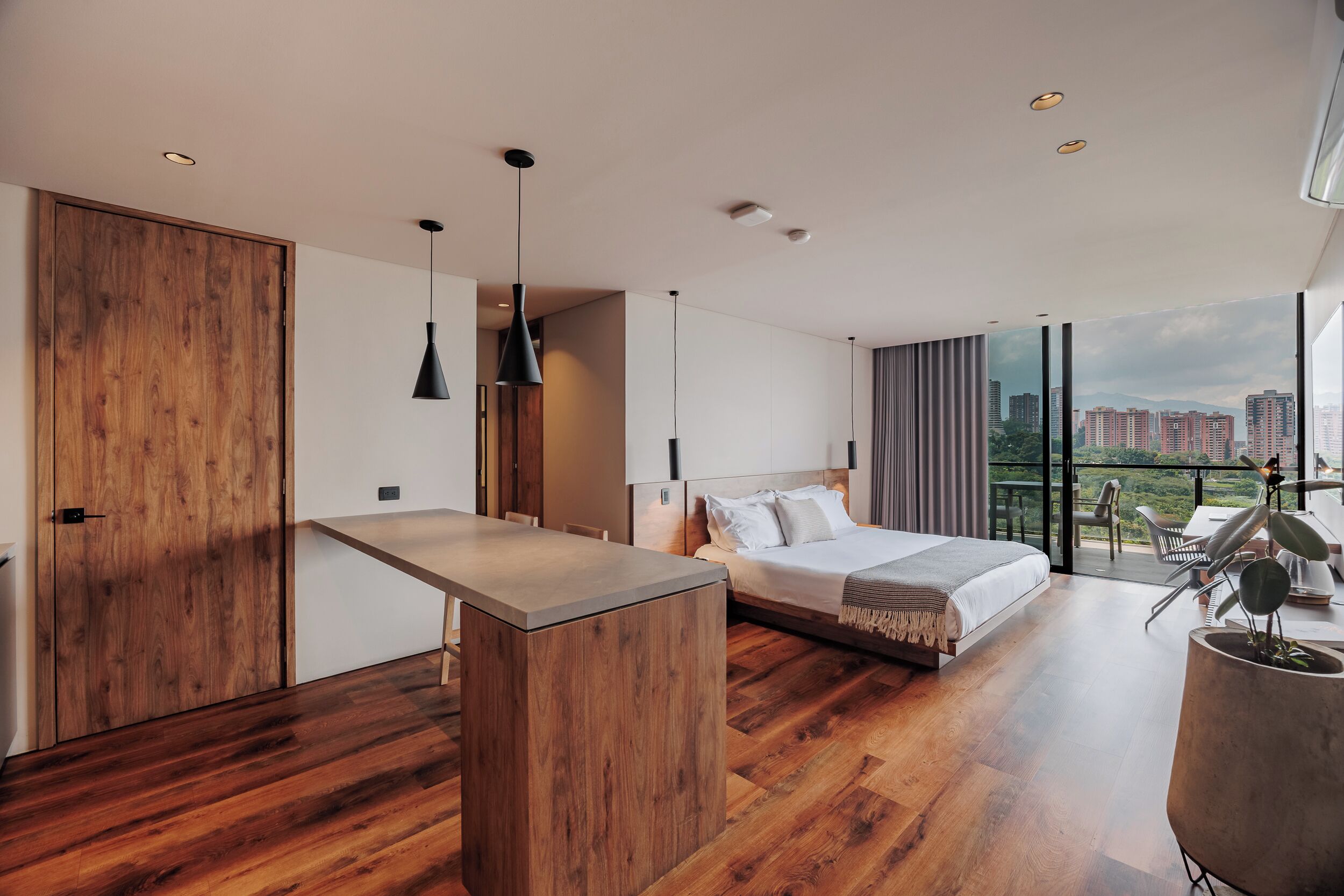 A modern bedroom with a large bed, a wooden desk, and a balcony with a city view.