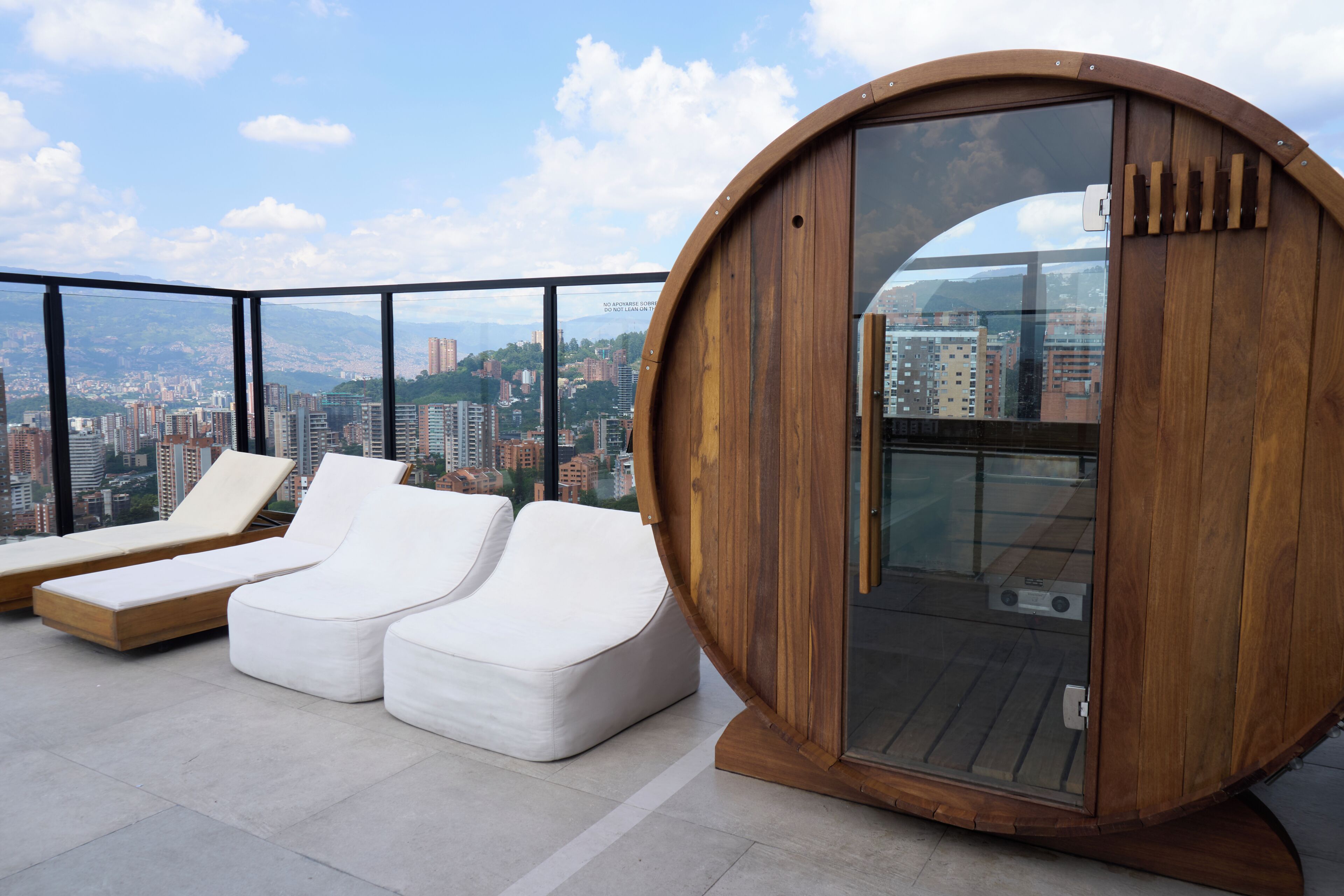 A rooftop lounge with white cushioned seating and a wooden structure with glass doors.