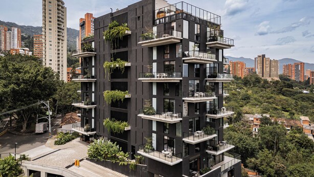 Un edificio a più piani con balconi, alcuni adornati con piante, circondato da altri edifici e vegetazione.