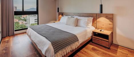 A modern hotel room with a large bed, a nightstand, a wooden headboard, a window with a city view, and an air conditioning unit.