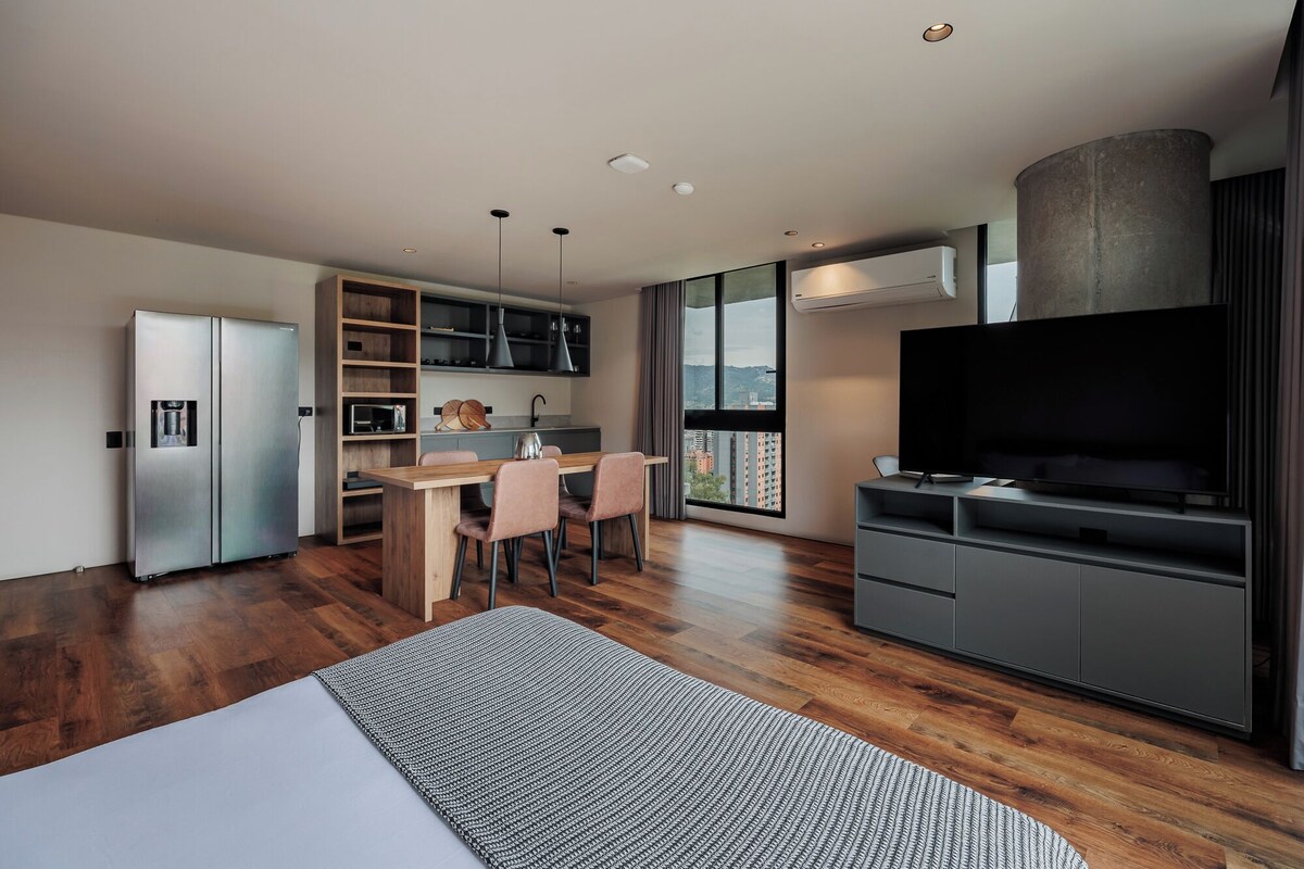 Una cucina moderna con frigorifero integrato, un'area pranzo con tavolo e sedie in legno, un'ampia finestra vista città e un televisore a schermo piatto su un mobile.
