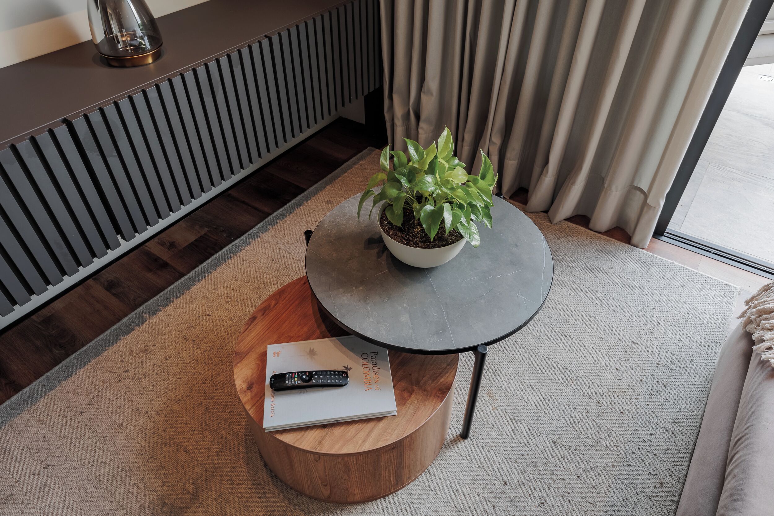 A modern living room with a round coffee table, a potted plant, a remote control, and a book on a wooden stool.
