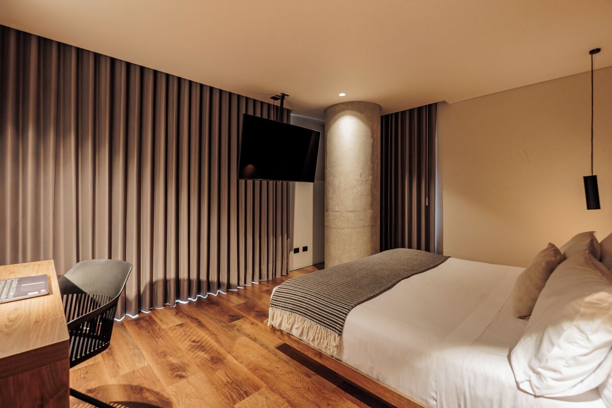 Una camera d'albergo moderna con pavimento in legno, un letto con lenzuola bianche, una scrivania con un portatile, una TV montata a parete e un'imponente colonna cilindrica.