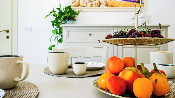 Una mesa de comedor con una bandeja de frutas, dos tazas blancas y un cuadro al fondo.