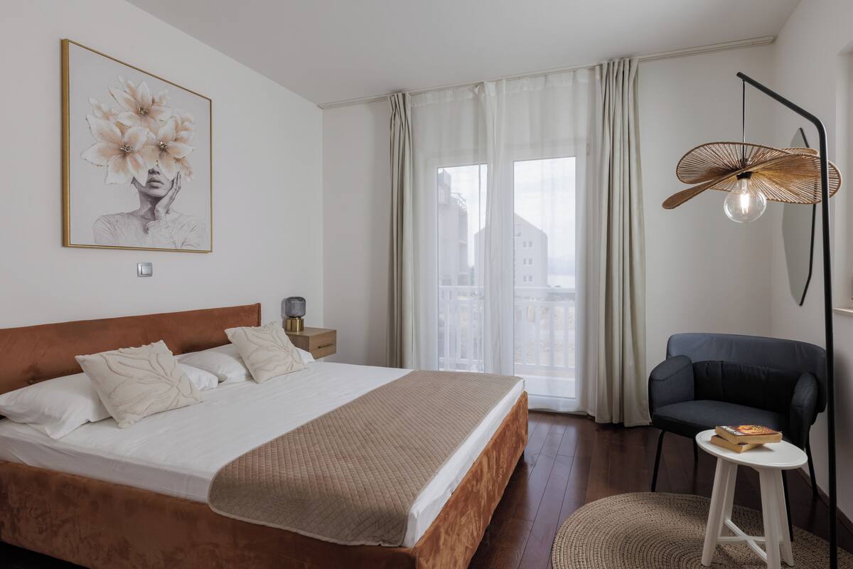 Un dormitorio con cama, una silla, mesita de noche, lámpara y ventana con cortinas.