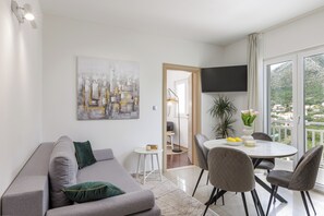 Ein modernes Wohnzimmer mit einem grauen Sofa, einem runden Esstisch mit Stühlen, einem flachbildfernseher an der Wand und einem großen Fenster mit Blick auf die Stadt.