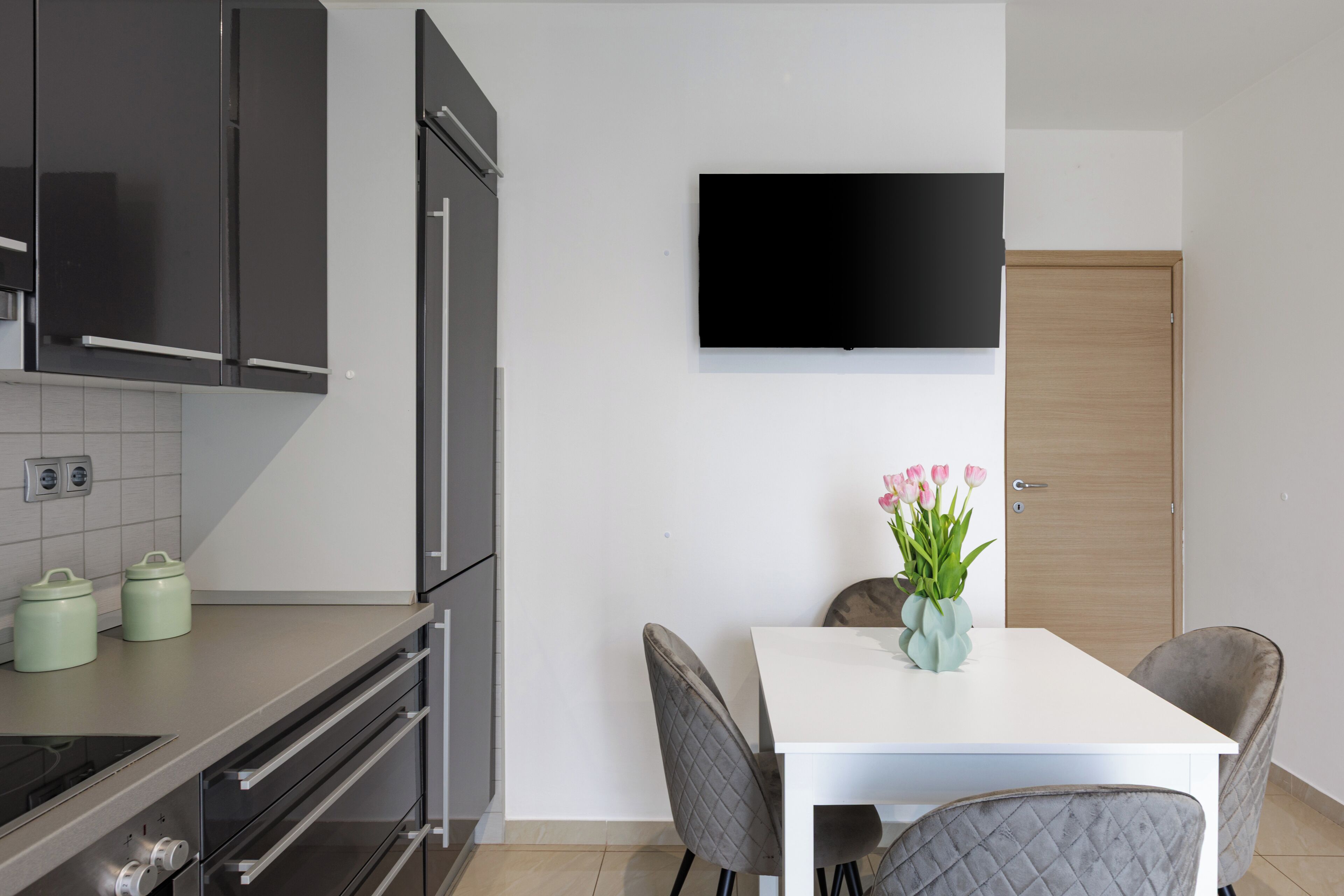 A modern kitchen with a flat-screen TV, a dining table with chairs, and a wooden door.