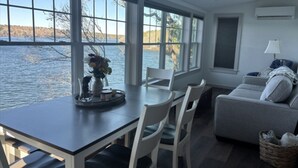Een kamer met een eettafel, stoelen, een bank en grote ramen met uitzicht op een wateroppervlak.