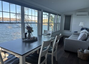 Ein Zimmer mit Esstisch, Stühlen, einer Couch und großen Fenstern mit Blick auf ein Gewässer.