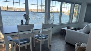 Een kamer met een eettafel, twee stoelen, een bank, een klein bijzettafeltje en uitzicht op een wateroppervlak door grote ramen.