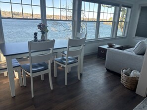 Ein Zimmer mit Esstisch, zwei Stühlen, einer Couch, einem kleinen Beistelltisch und Blick auf ein Gewässer durch große Fenster.