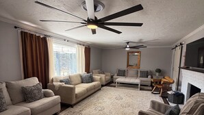 Ein Wohnzimmer mit Deckenventilator, Kamin und großem Fenster mit Vorhängen.