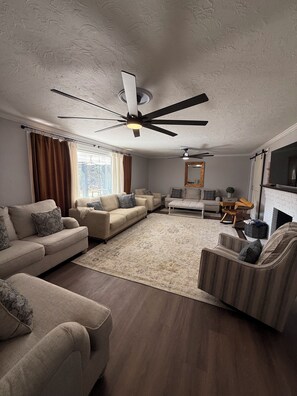 Ein Wohnzimmer mit Deckenventilator, Kamin und großem Fenster mit Vorhängen.