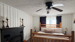 Ein Schlafzimmer mit Bett, Kamin, Deckenventilator und einem Fenster mit Vorhängen.