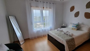 A bedroom with a bed, a bedside table with a lamp, a window with sheer curtains, and a wall with decorative leaf patterns.