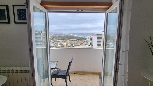 A balcony with a view of the sea and buildings, a chair, a table, and a potted plant.
