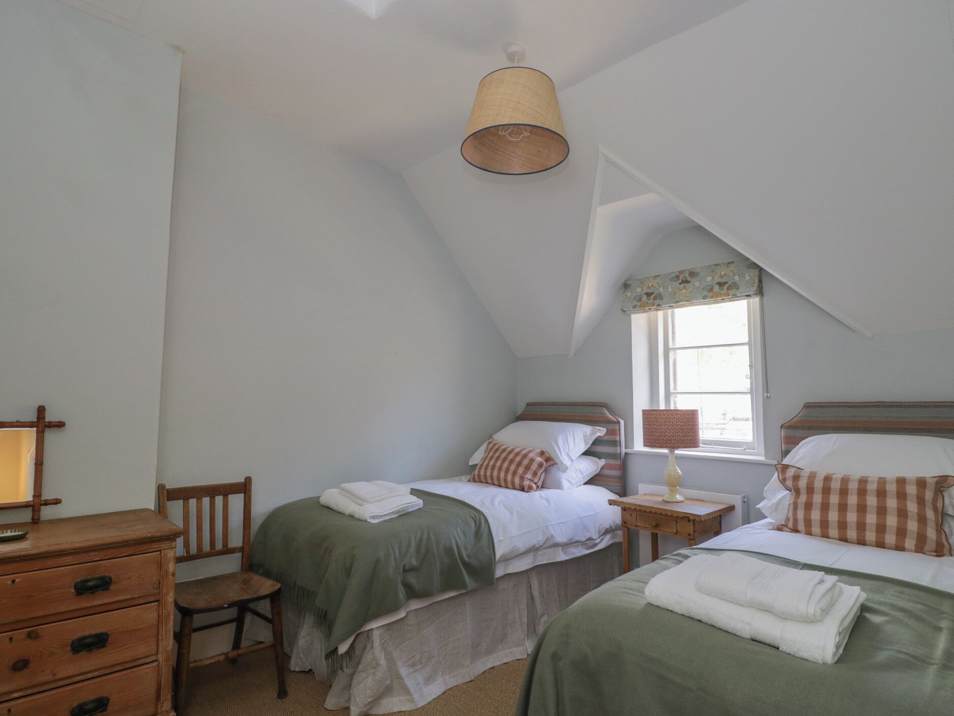 Une chambre avec deux lits, une commode en bois, une chaise, une lampe et une fenêtre avec un rideau.
