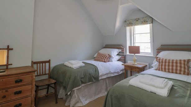 A bedroom with two beds, a wooden dresser, a chair, a lamp, and a window with a curtain. - Middle Cottage (Salisbury)