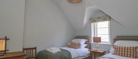 Une chambre avec deux lits, une commode en bois, une chaise, une lampe et une fenêtre avec un rideau.