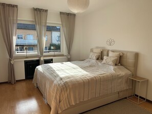 Ein Schlafzimmer mit Bett, einem Fenster mit Vorhängen, einem Heizkörper, einem Nachttisch und einer Wanddekoration.