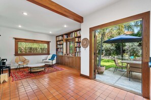 Ein geräumiger Raum mit großem Fenster, einer Bücherwand, einem Essbereich mit Sonnenschirm und einer gemütlichen Sitzecke.